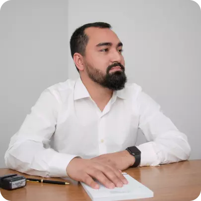 Homem sentado por trás de uma mesa. Ele está com o olhar direcionado para a direita. Suas mãos estão sobre um caderno.