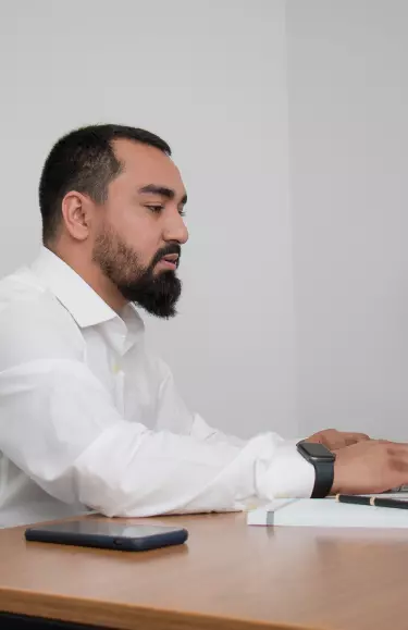 Homem sentado em frente a uma mesa mexendo em um notebook. Ele usa uma camisa social e um relógio em seu pulso.