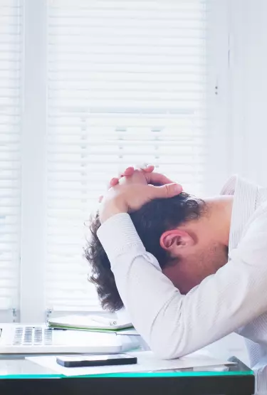 Homem sentado em frente a uma mesa. Ele está com a cabeça baixa e com as duas mãos sobre a nuca.