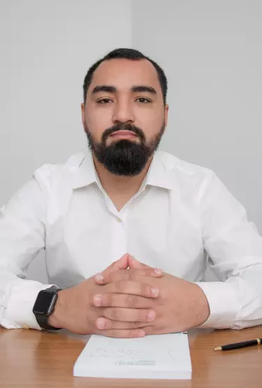 Homem sentado em frente a uma mesa mexendo em um notebook. Ele usa uma camisa social e um relógio em seu pulso.