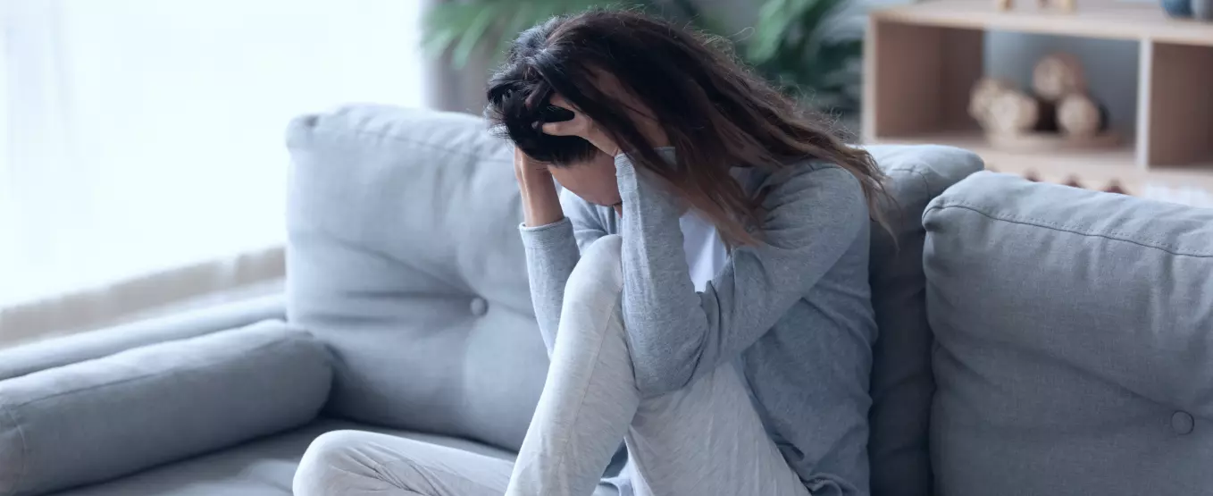 Mulher sentada em um sofá com umas das pernas sobre o móvel. Ela está com os cabelos soltos e cabeça baixa. As suas duas mãos estão sobre o início da sua cabeça.